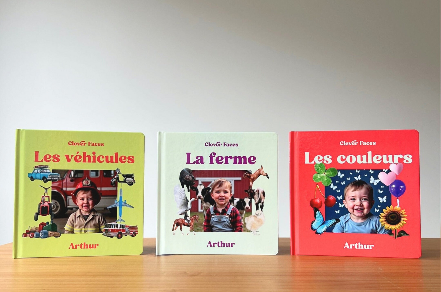 Three personalized children's hardcover books from Clever Faces are lined up on a wooden surface against a neutral gray background. The books are titled 'Les véhicules' in yellow, 'La ferme' in off-white, and 'Les couleurs' in red, each featuring a custom photo of a child and their name.
